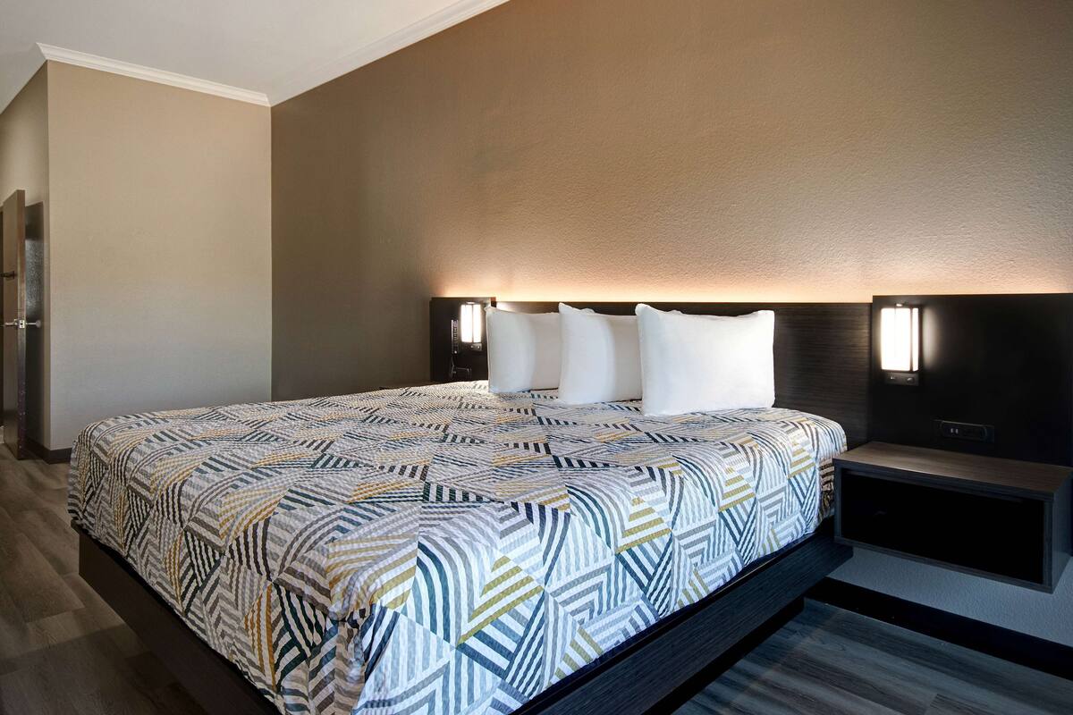 A modern hotel room with a large bed, a headboard with built-in lighting, and a bedside table.