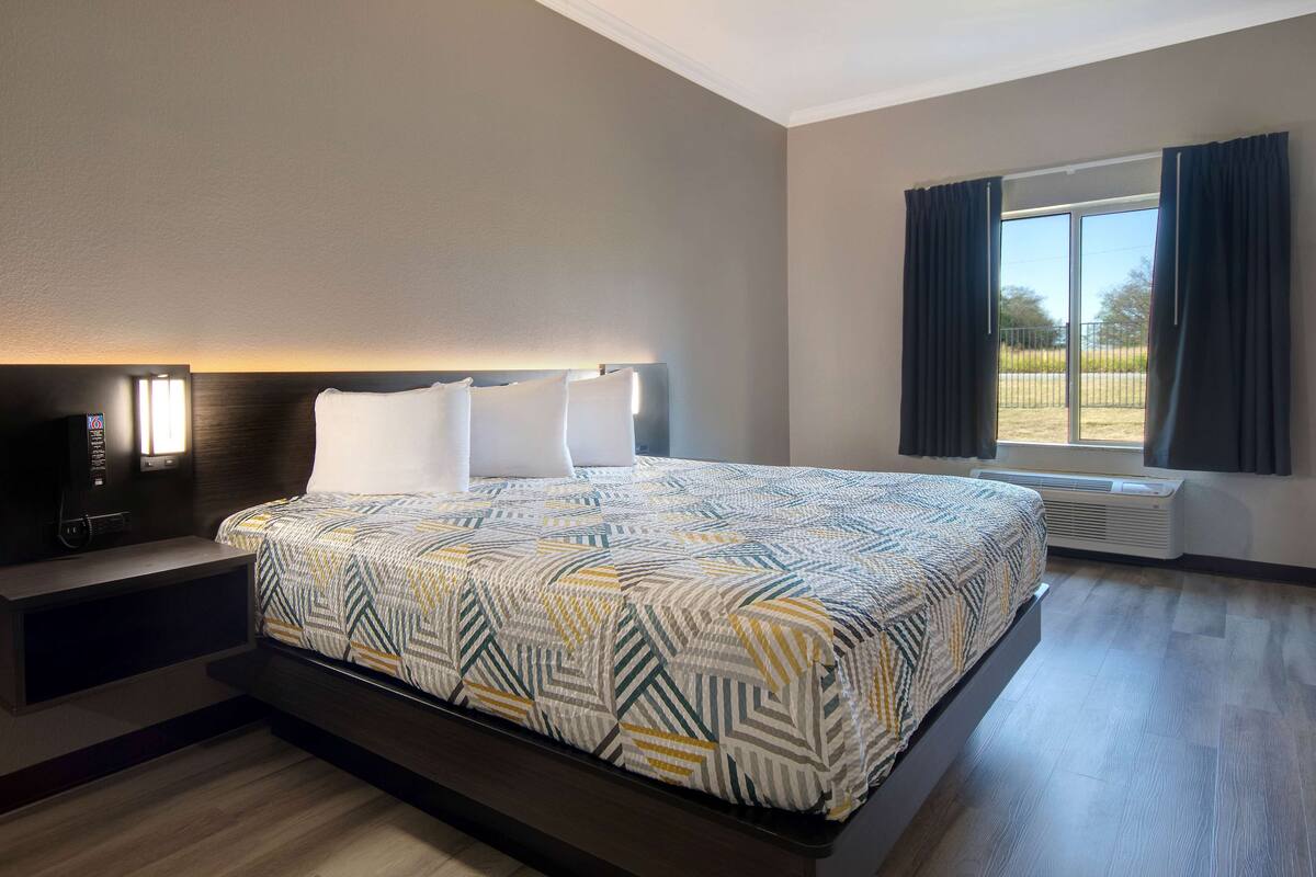 A hotel room with a large bed, a bedside table with a lamp, a window with curtains, and a view of the outdoors.