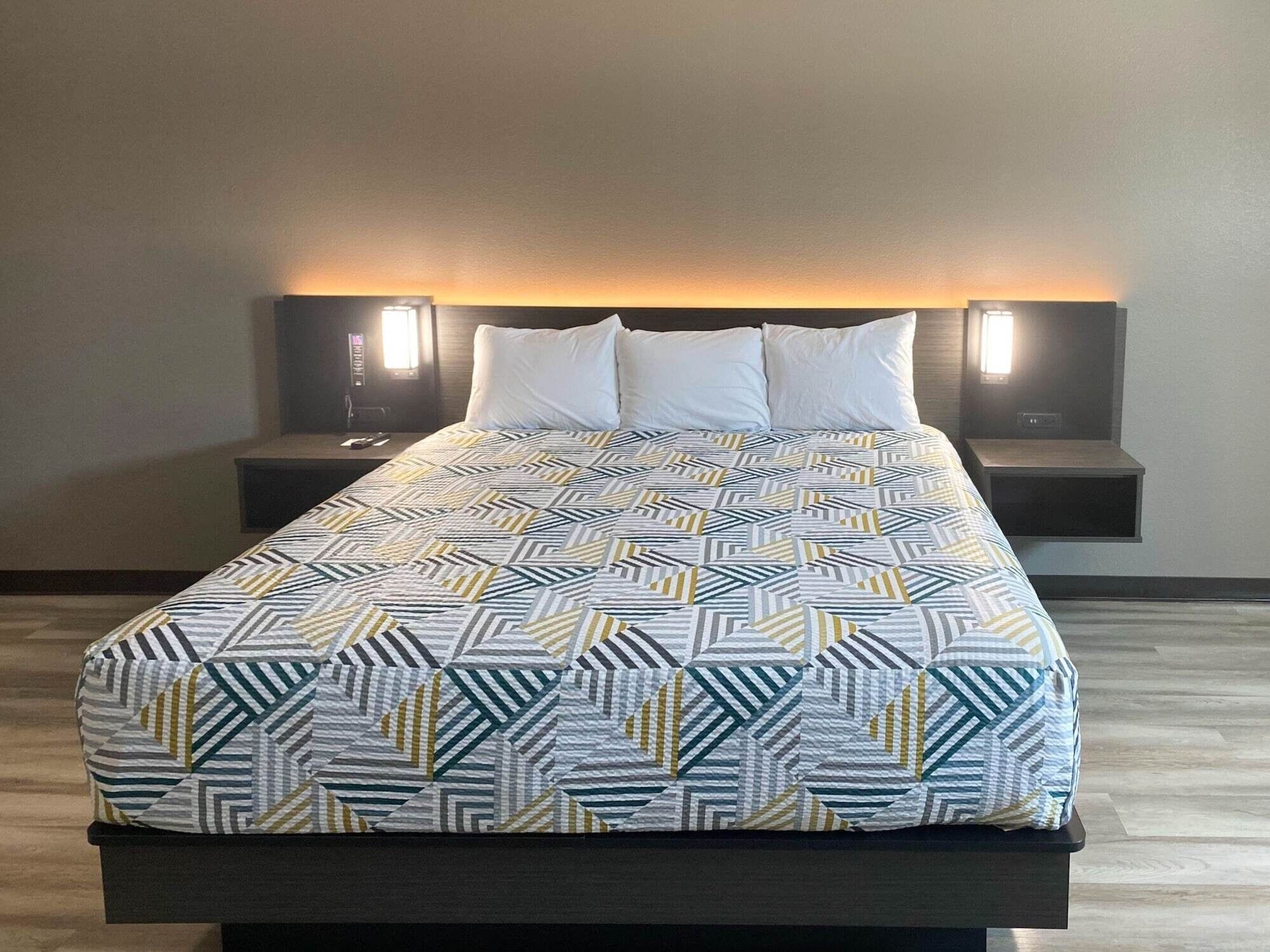 Una cama bien hecha con una colcha estampada, dos mesitas de noche con lámparas, y suelo de madera.