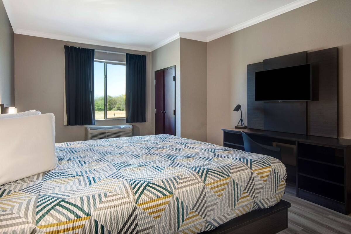 A hotel room with a large bed, a flat-screen TV mounted on the wall, a desk with a chair, and a window with curtains.