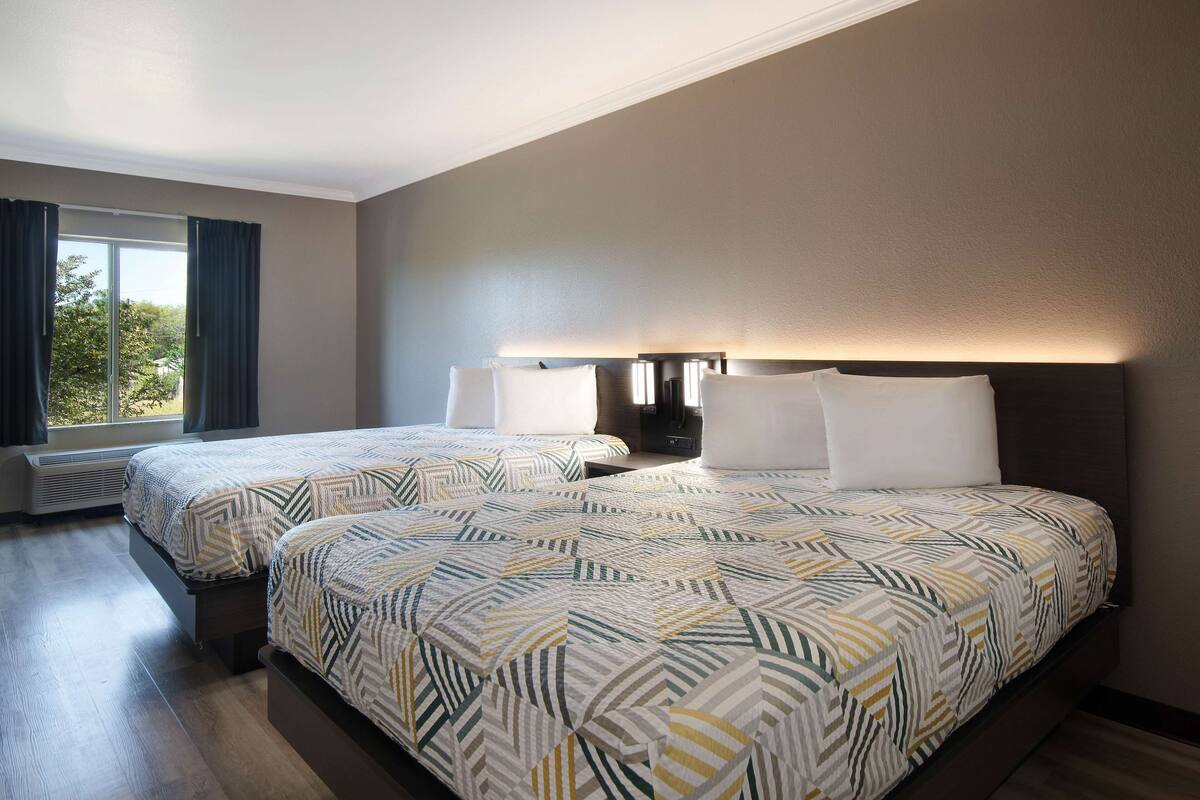 A hotel room with two beds, a large window with curtains, a wall-mounted lamp, and a built-in heater.