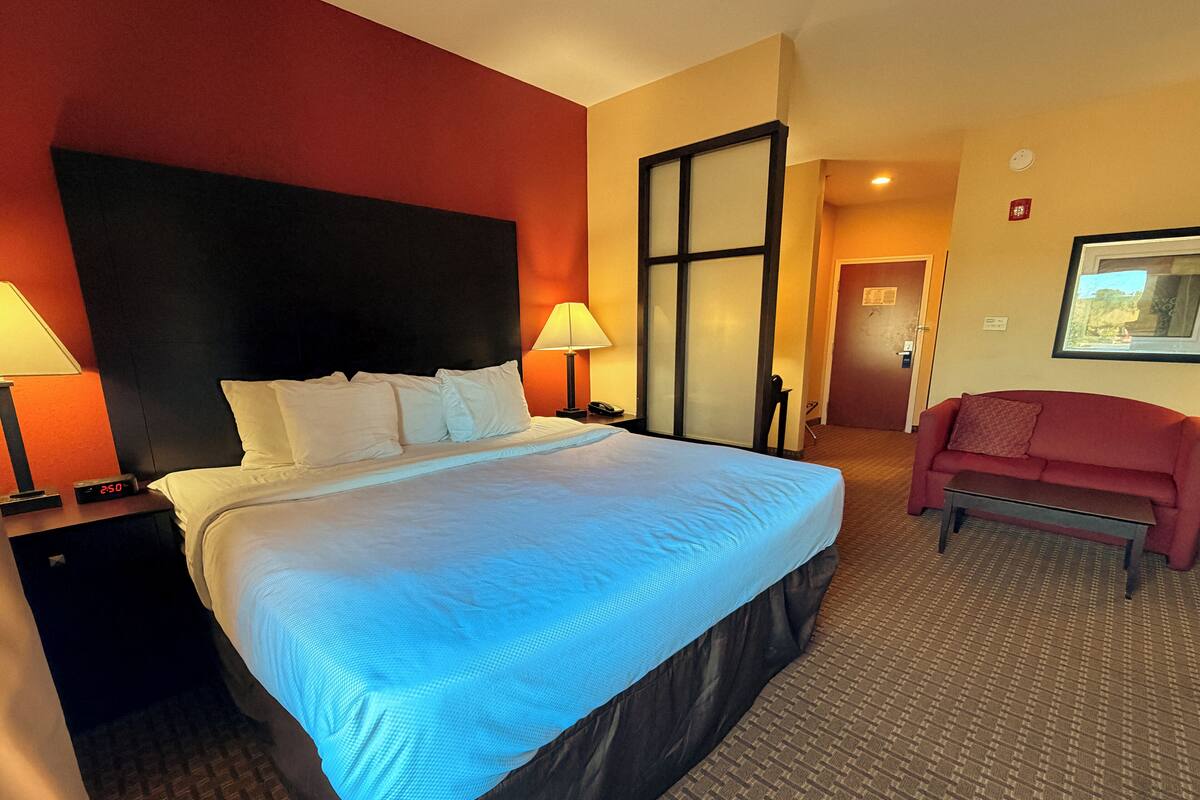 A hotel room with a large bed, two bedside tables with lamps, a red sofa, and a door leading to another room.