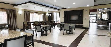A spacious dining area with multiple tables and chairs, a large flat-screen TV, and a kitchen area in the background.
