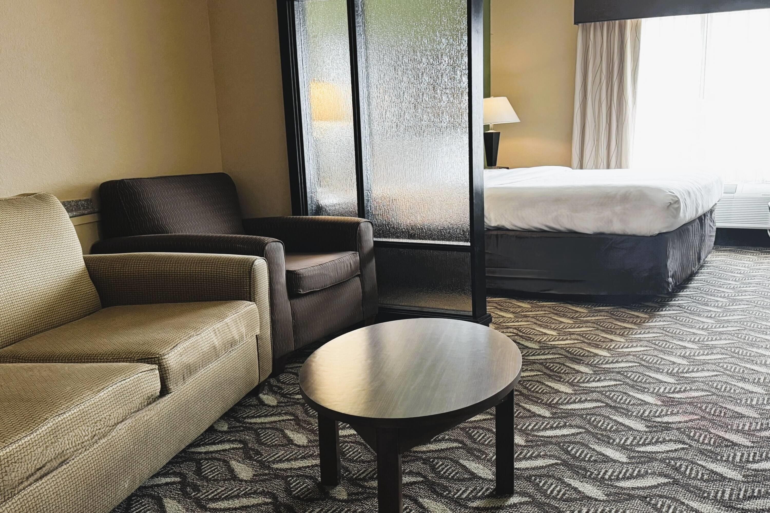 A hotel room with a sofa, armchair, and a small table. There is a bed with a white comforter and a lamp on the nightstand.