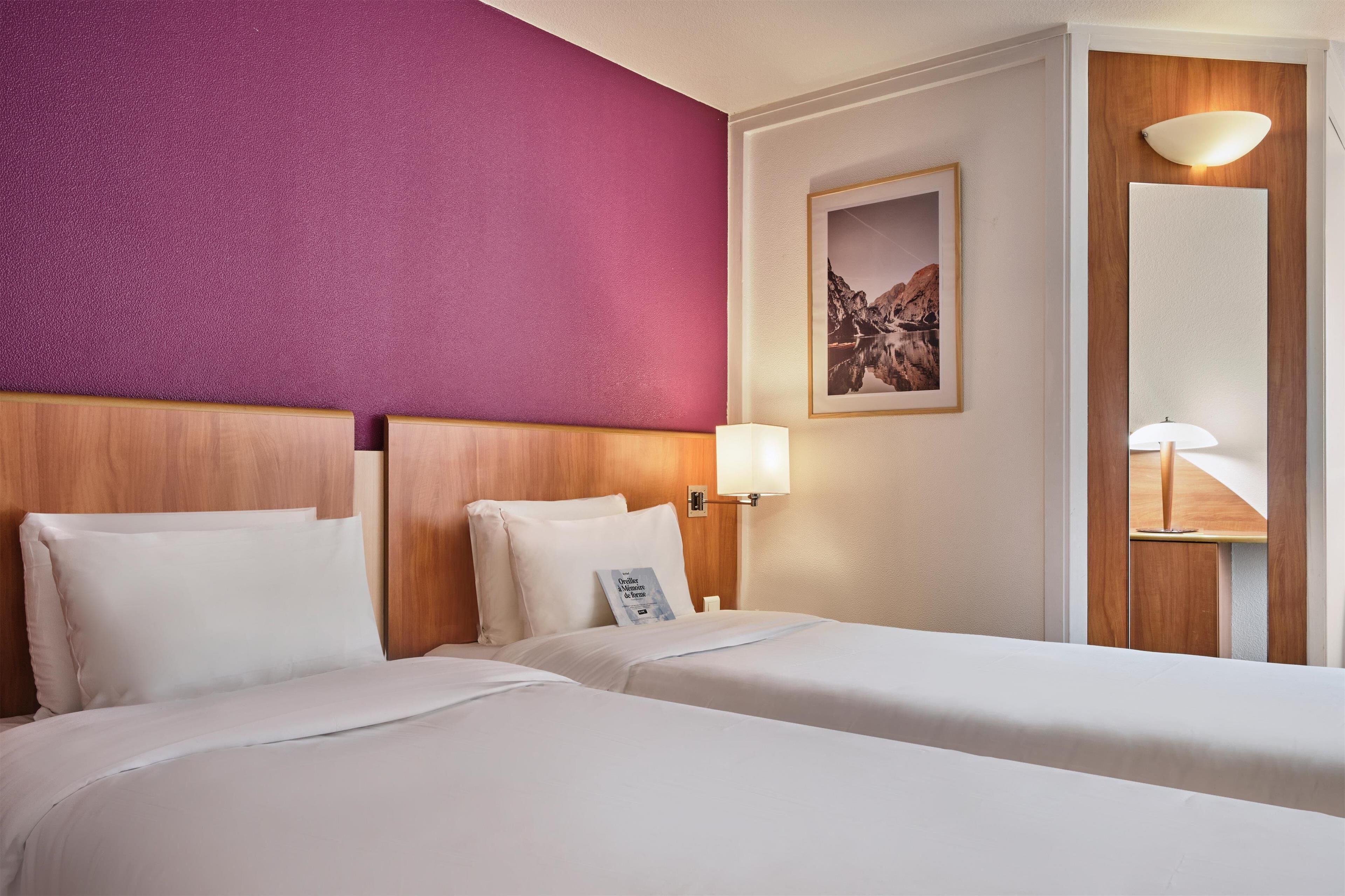 Habitación de hotel con dos camas, cabecera de madera, pared de acento púrpura, un cuadro enmarcado y una lámpara de noche.