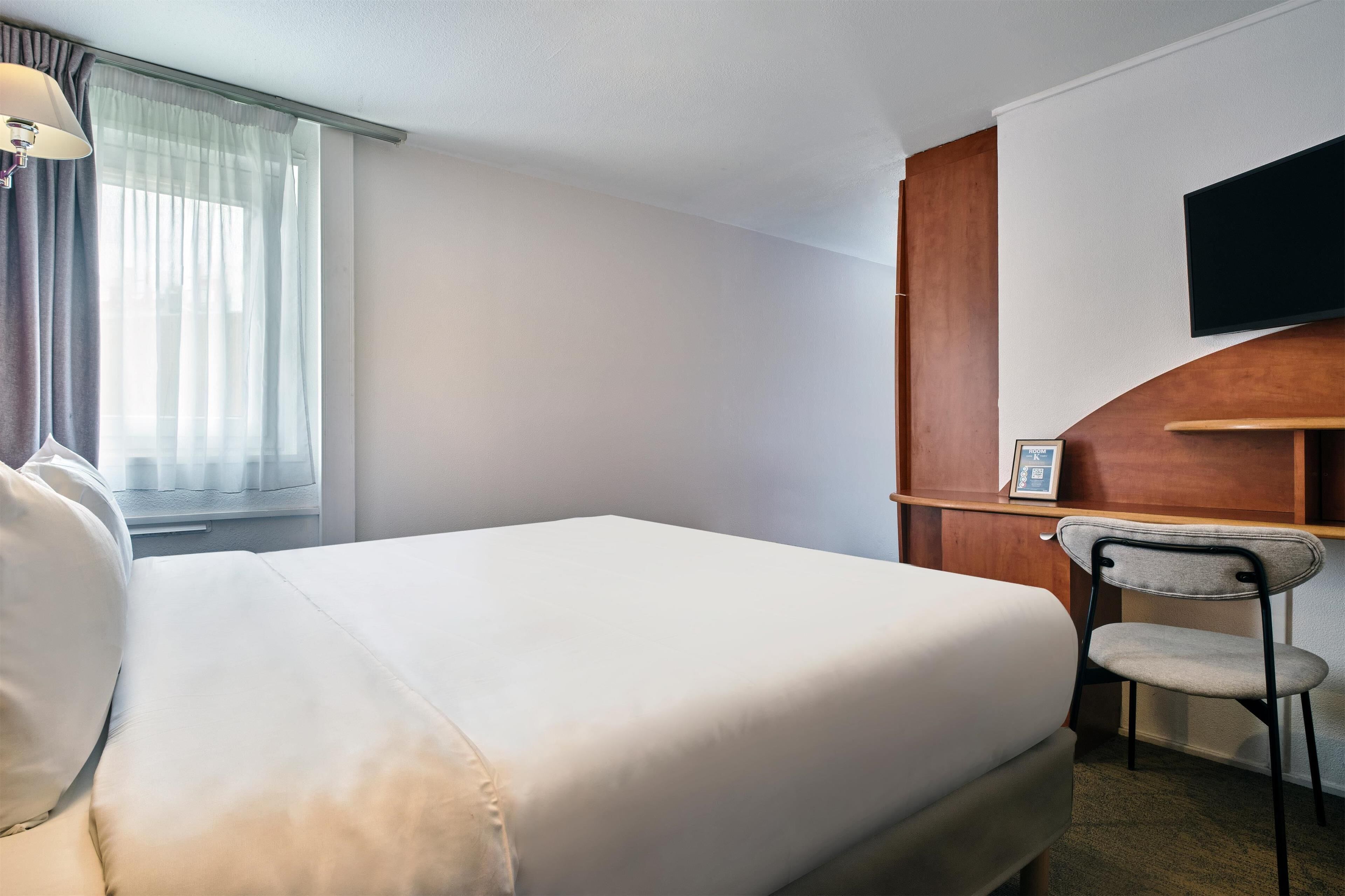 A hotel room with a bed, a desk, a chair, a TV mounted on the wall, and a window with curtains.