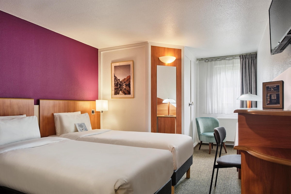 A hotel room with two beds, a desk, a chair, a lamp, and a framed picture on the wall.