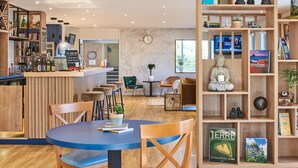 A modern bar area with a counter, stools, and a variety of bottles. There is a seating area with a table and chairs, and a bookshelf with books and decorative items. - Kyriad Les Ulis (Les Ulis)