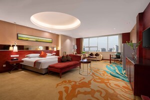 Habitación de hotel con cama, sofá rojo, mesita, televisor y ventana grande con vista a la ciudad.