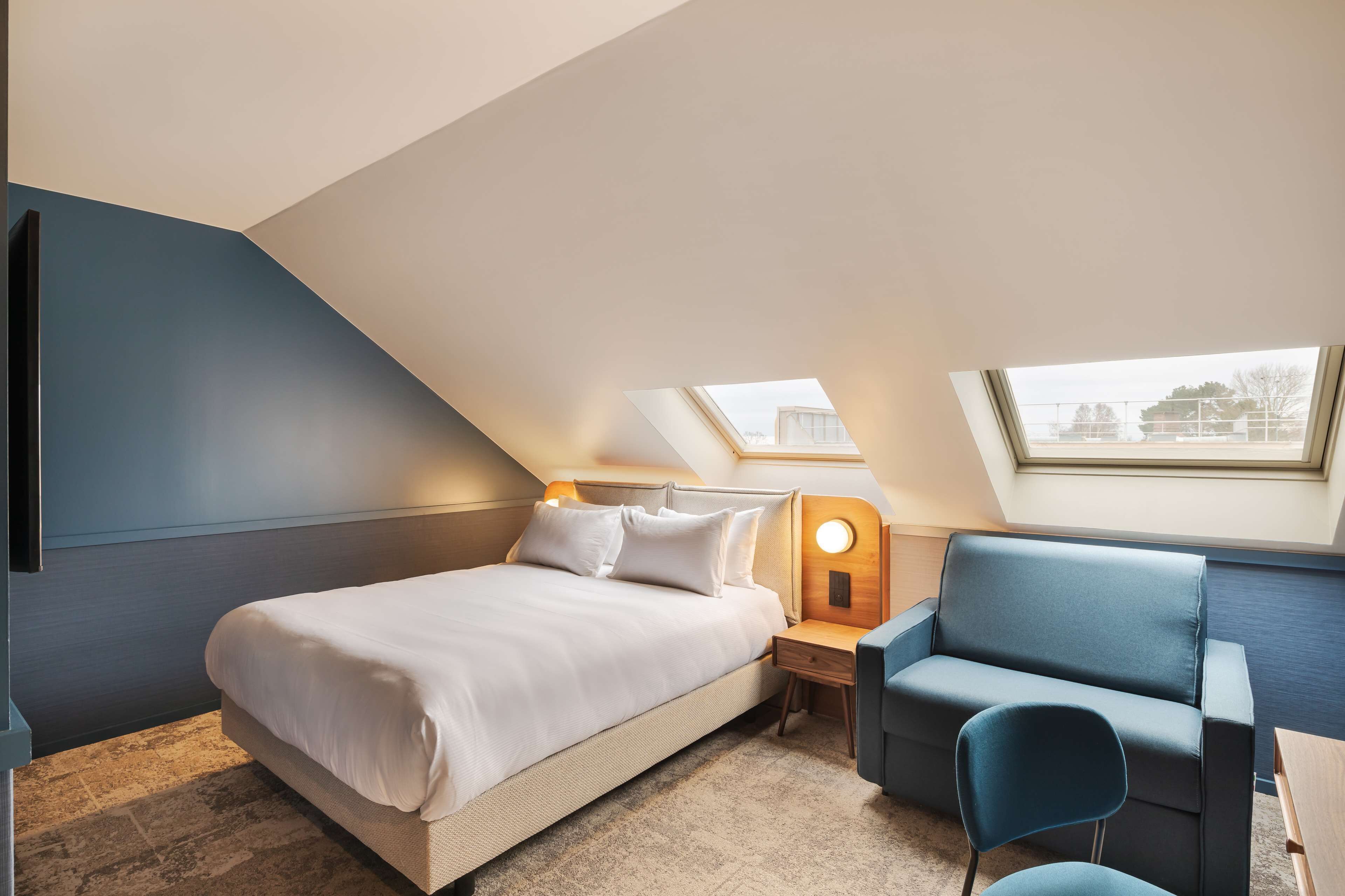 A modern hotel room with a bed, a desk, a chair, and a skylight.