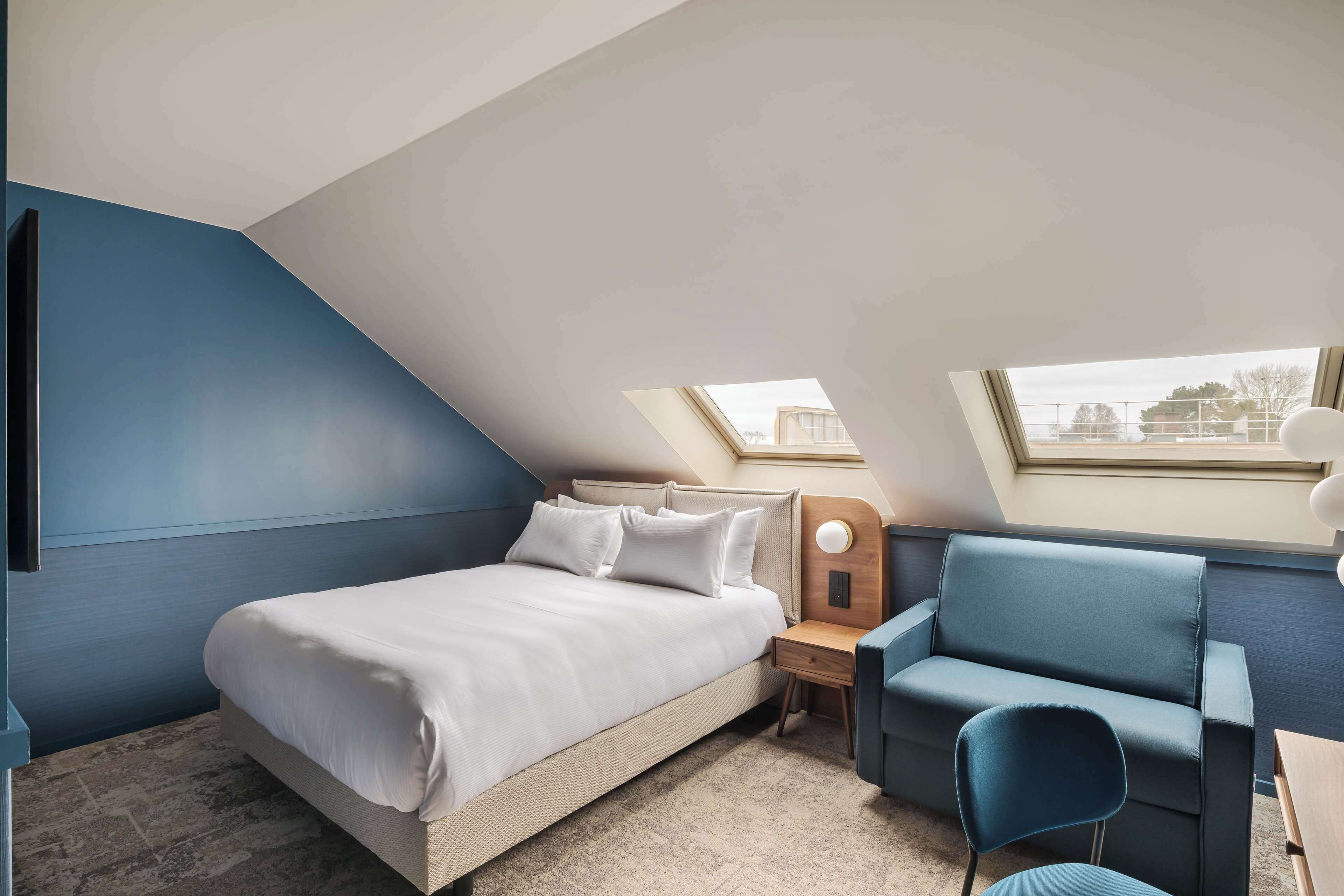 A bedroom with a bed, a nightstand, a blue chair, and a desk.