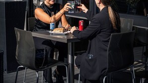 Twee personen zitten aan een buitentafel met een fles bier, een blikje fris en een bord eten.