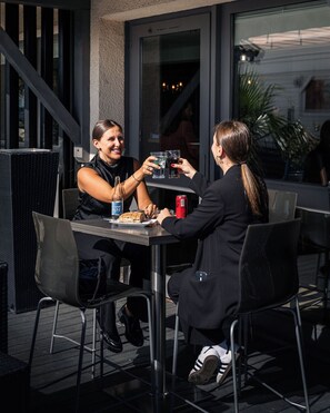 Dos personas sentadas en una mesa al aire libre con una botella de cerveza, una lata de refresco y un plato de comida.