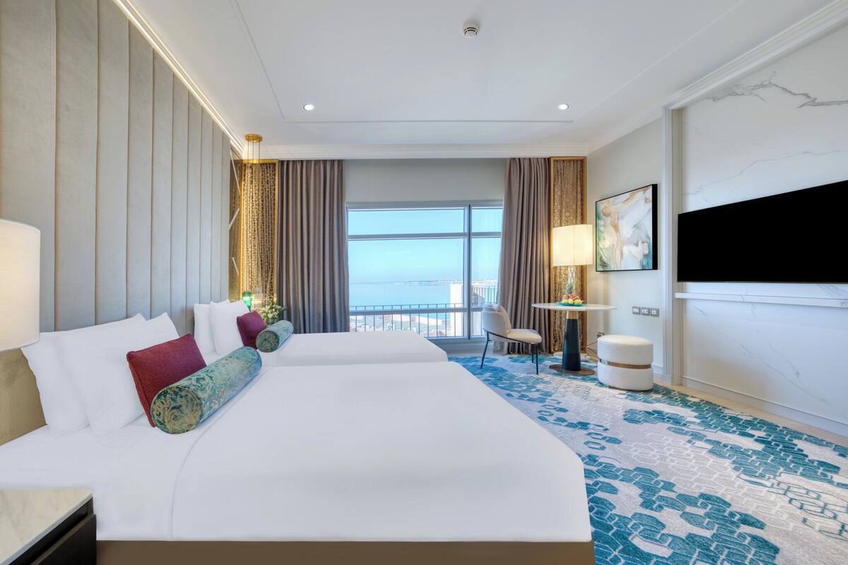 A hotel room with two beds, a large window with a view, a flat-screen TV, and a small table with a lamp.