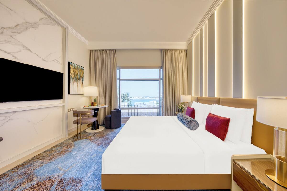 A hotel room with a large bed, a TV mounted on the wall, a desk with a chair, and a view of the city through the window.