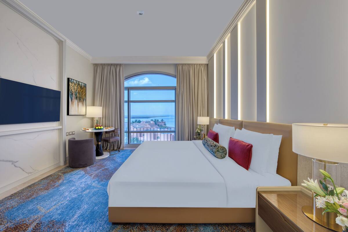 A hotel room with a large bed, a desk, a chair, a small table with fruit, and a view of the ocean through a large window.