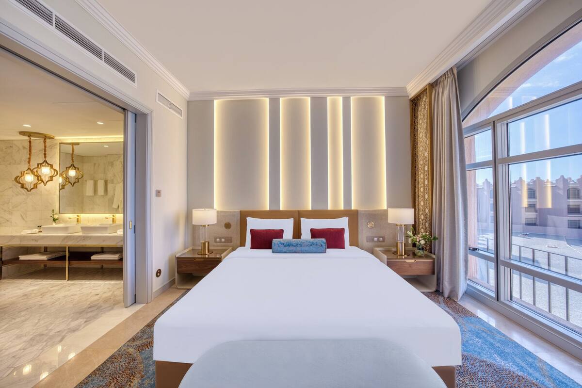 A hotel room with a large bed, a bathroom with a double sink, and a city view through the window.