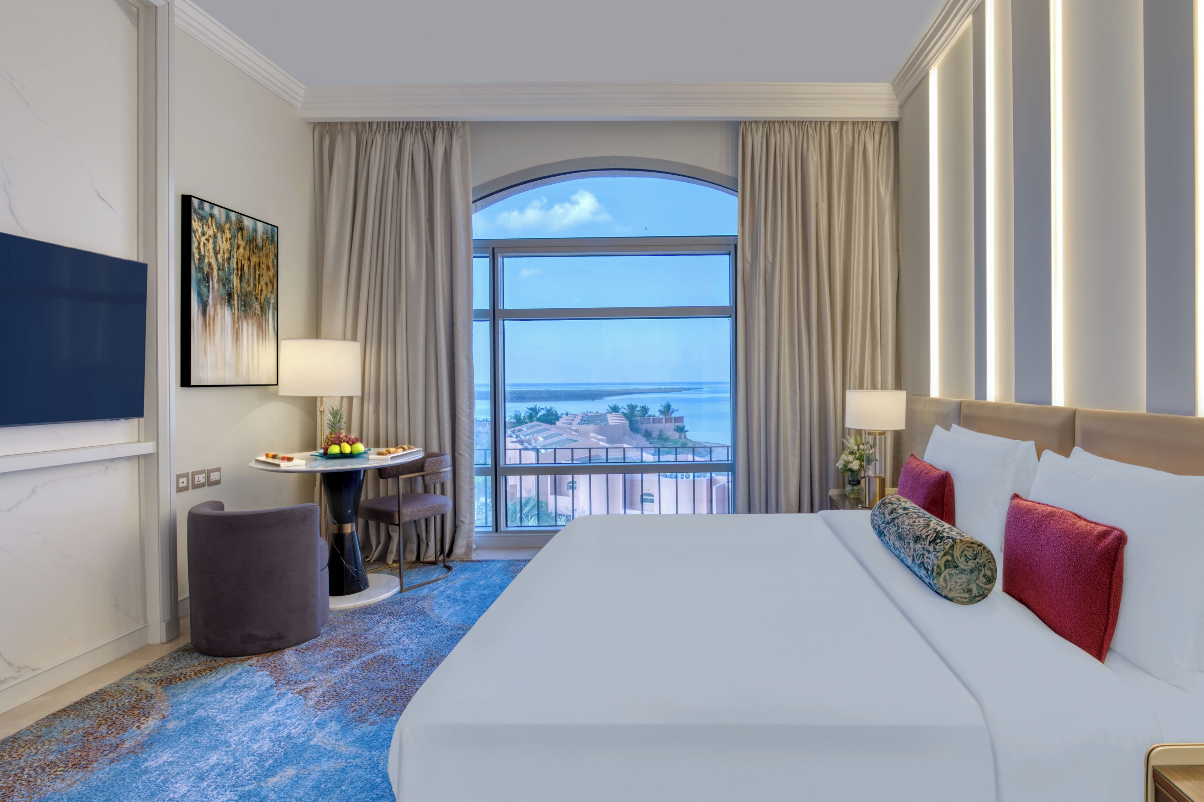 A hotel room with a large bed, a TV, a dining table with fruit, and a view of the sea.