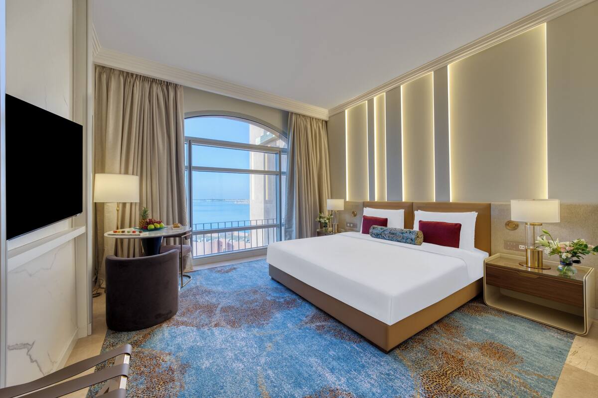 A hotel room with a large bed, a TV, a small table with fruit, a nightstand, and a view of the sea.