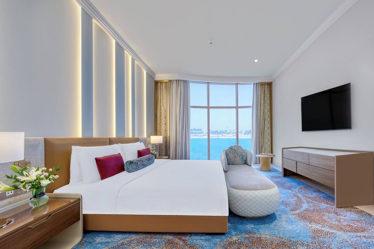 A hotel room with a large bed, a desk, a chair, a TV, and a view of the ocean.