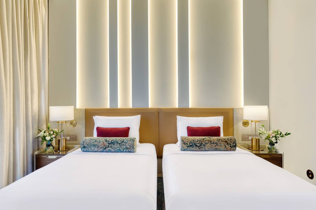 A hotel room with two beds, each with a red pillow, a blue patterned pillow, and a bedside table with a lamp and a vase of flowers.