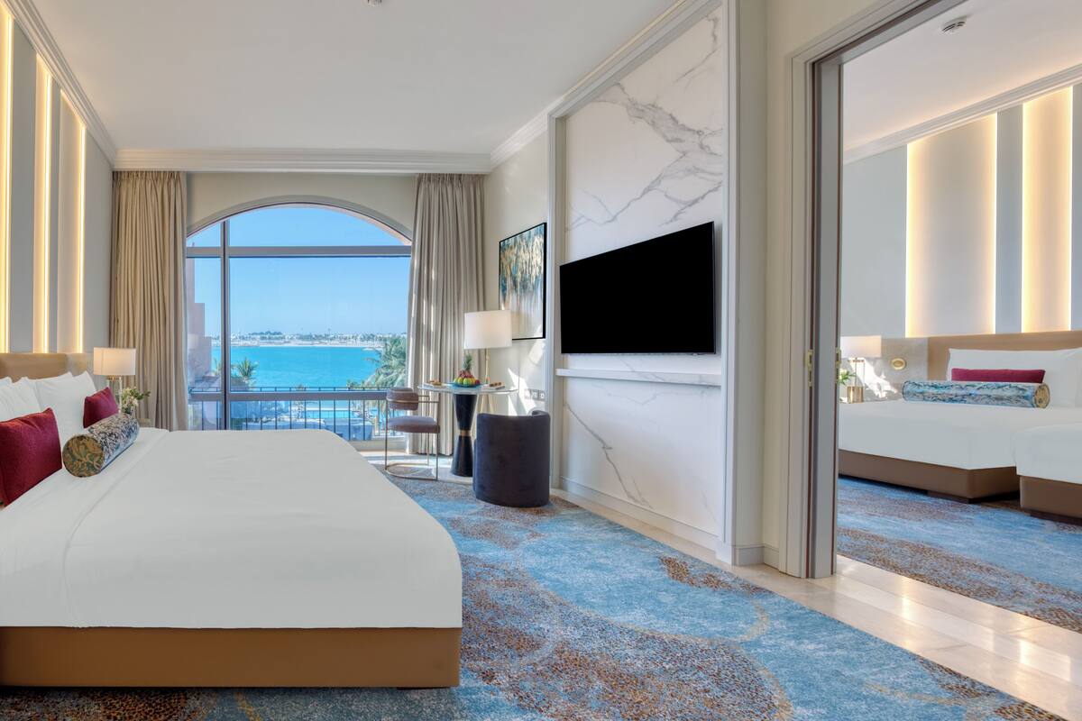 A hotel room with a large bed, a TV, a desk with a chair, and a view of the sea.