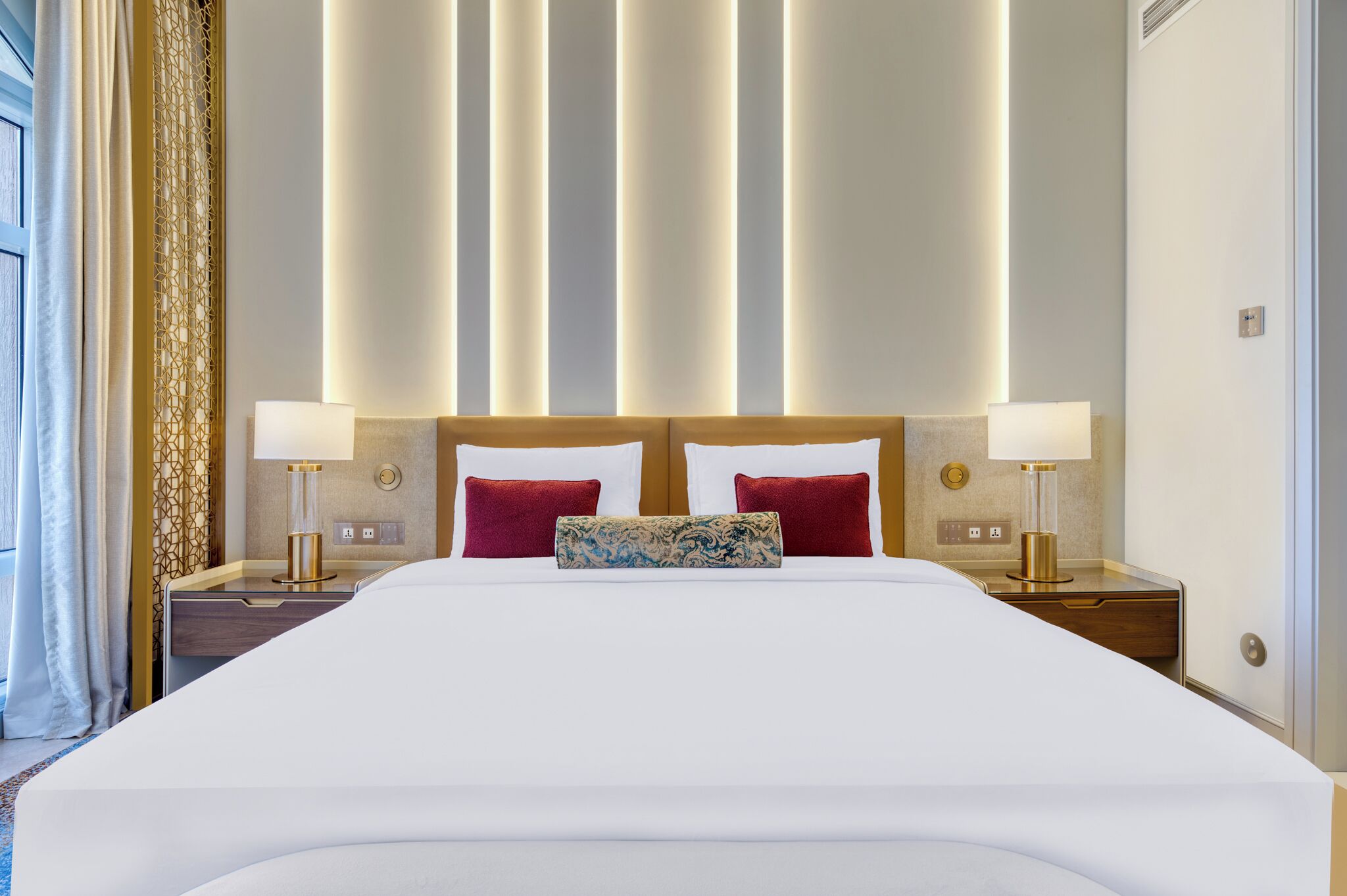 Un grand lit avec du linge de lit blanc, deux oreillers rouges et un coussin décoratif. Il y a deux tables de chevet avec des lampes, un panneau mural décoratif et une fenêtre avec des rideaux.