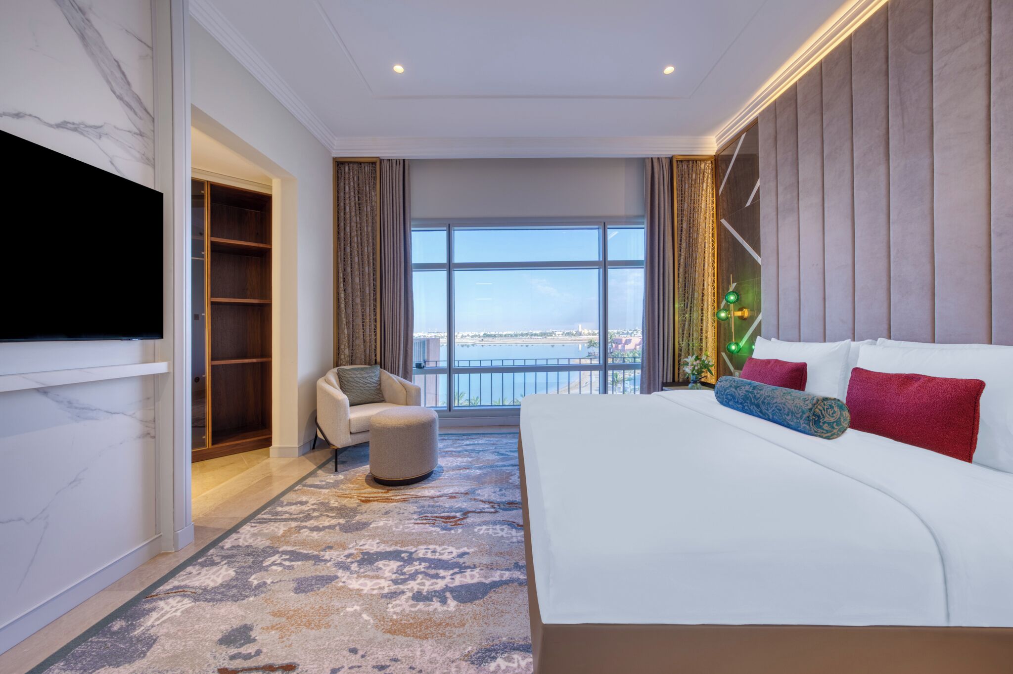 A modern hotel room with a large bed, a TV mounted on the wall, a seating area with a chair and ottoman, a built-in bookshelf, and a large window offering a city view.