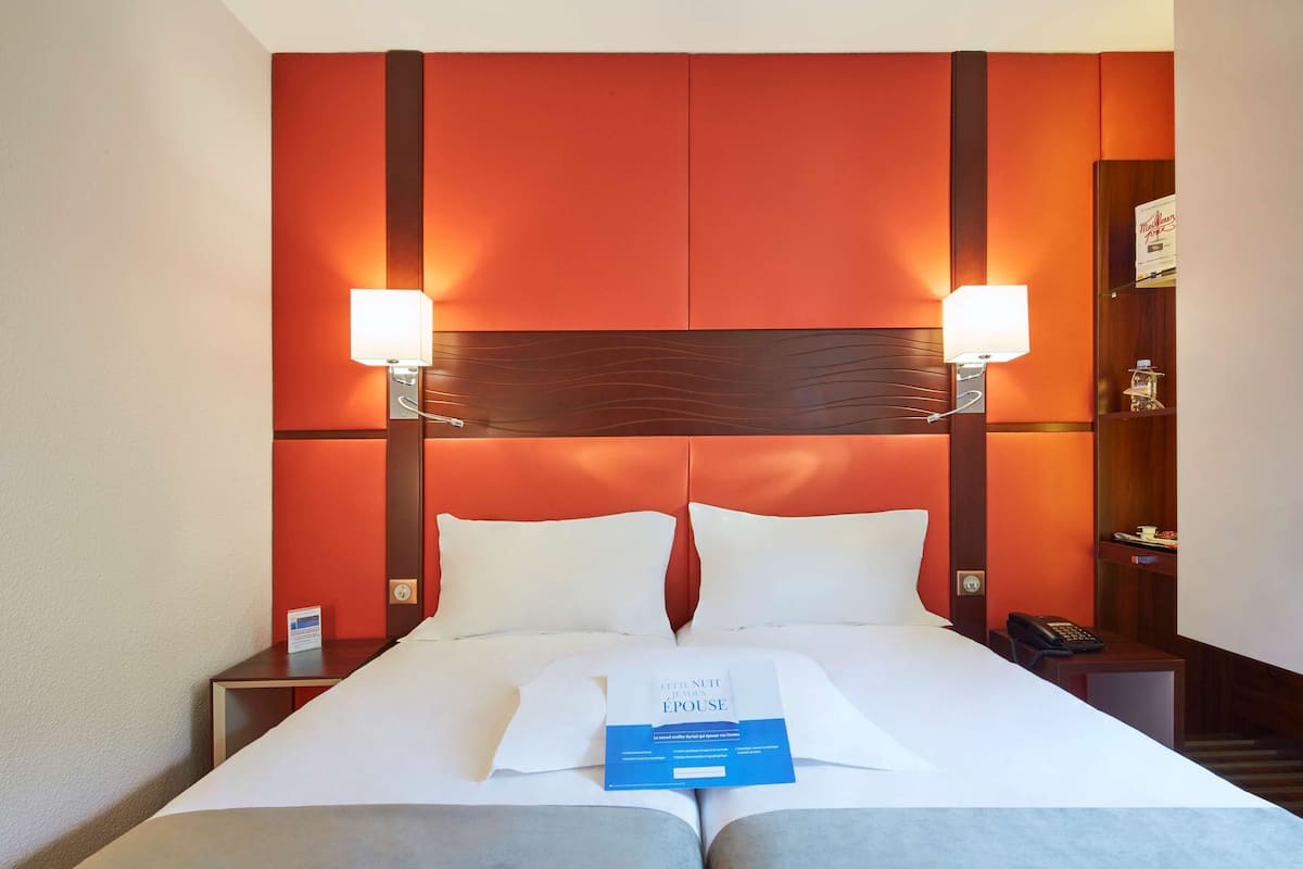A hotel room with a large bed, two bedside lamps, a nightstand with a phone, and a small shelf with a bottle and a sign.