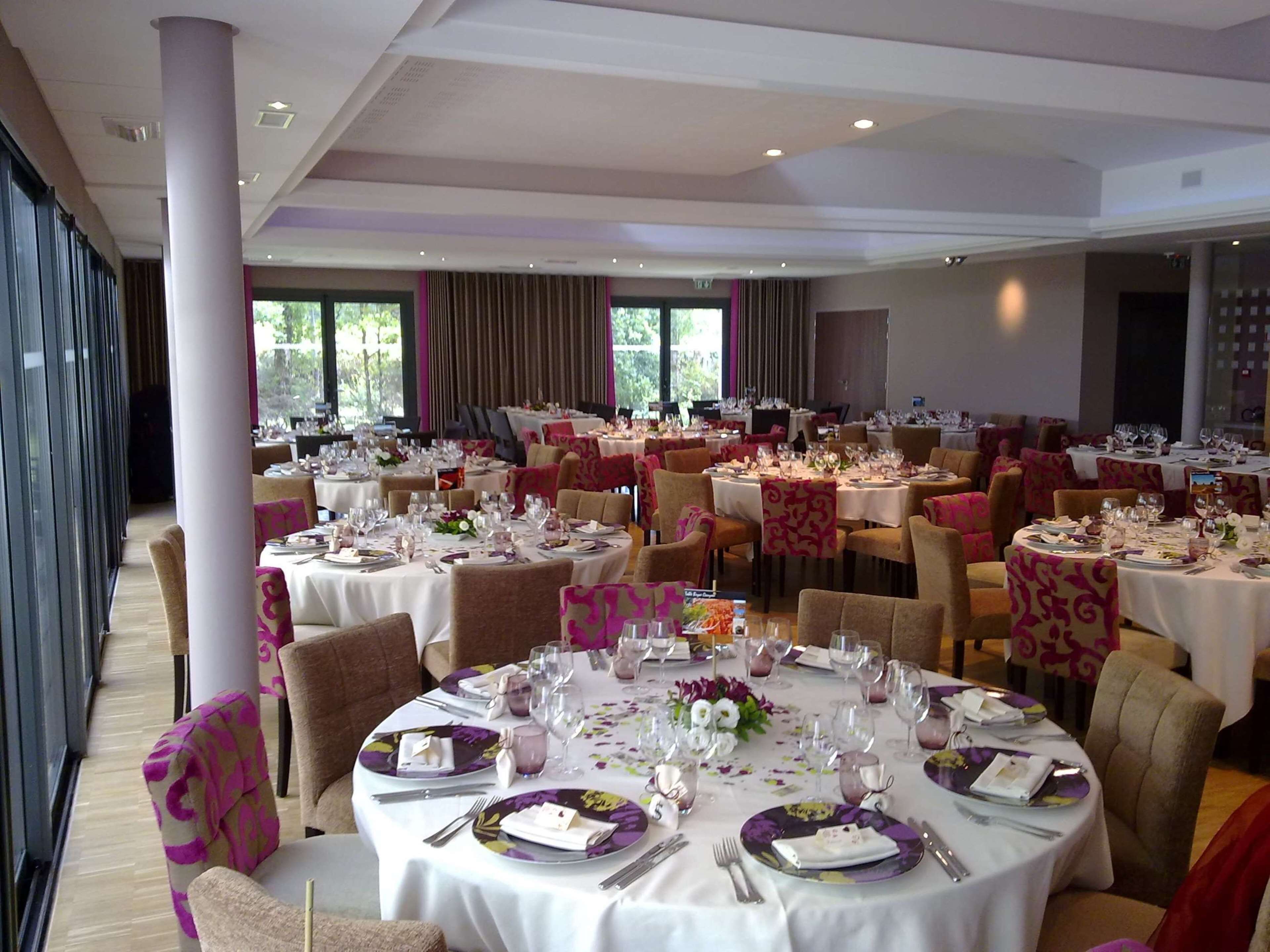 Une salle de banquet avec de nombreuses tables rondes dressées pour le repas, agrémentées de nappes blanches et de compositions florales violettes.