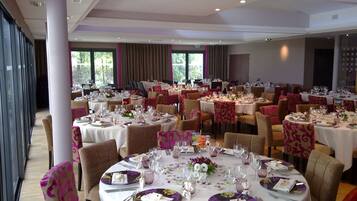 A banquet hall with multiple round tables set for dining, featuring white tablecloths and purple floral centerpieces.