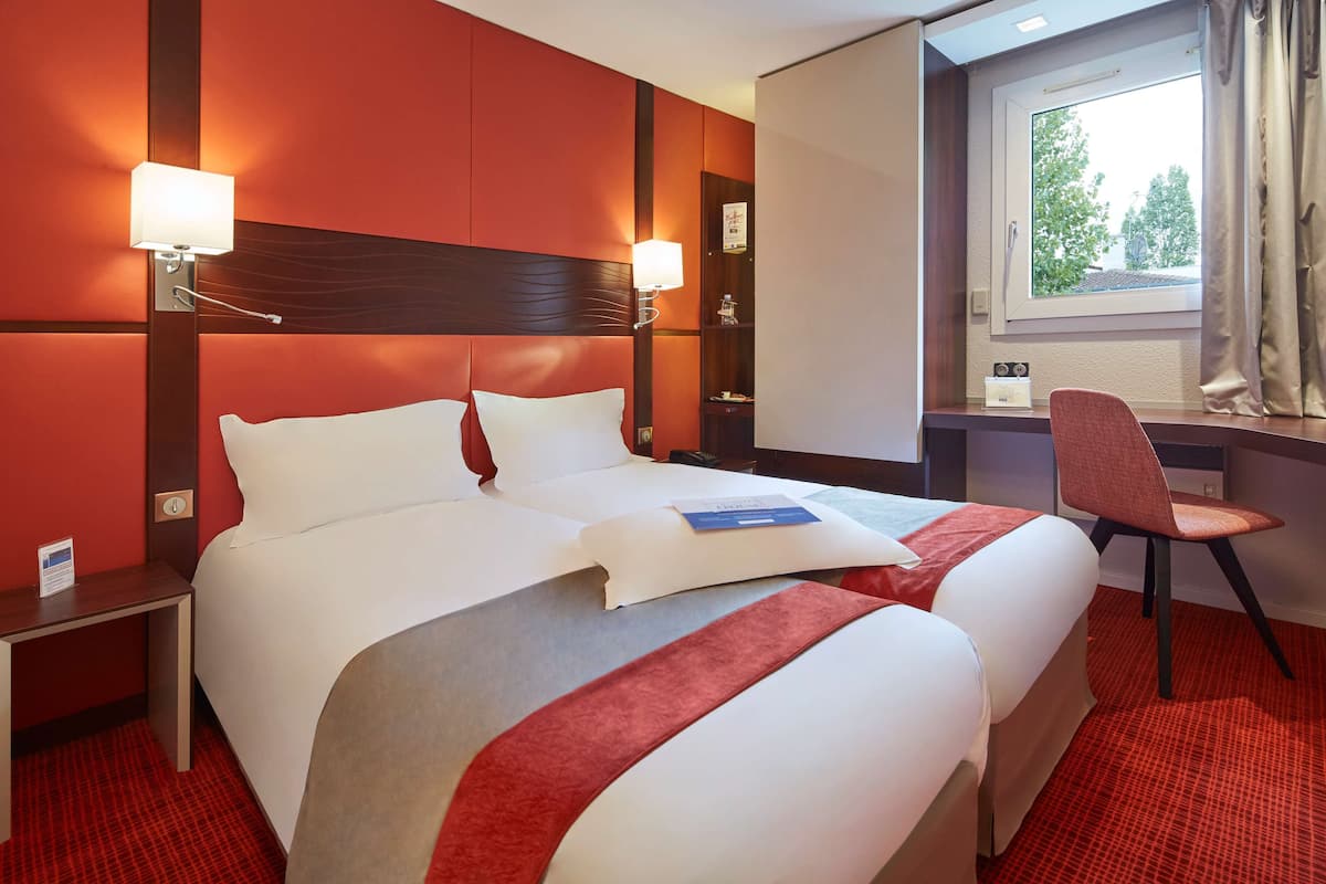 A hotel room with a large bed, two bedside lamps, a desk with a chair, and a window with curtains.