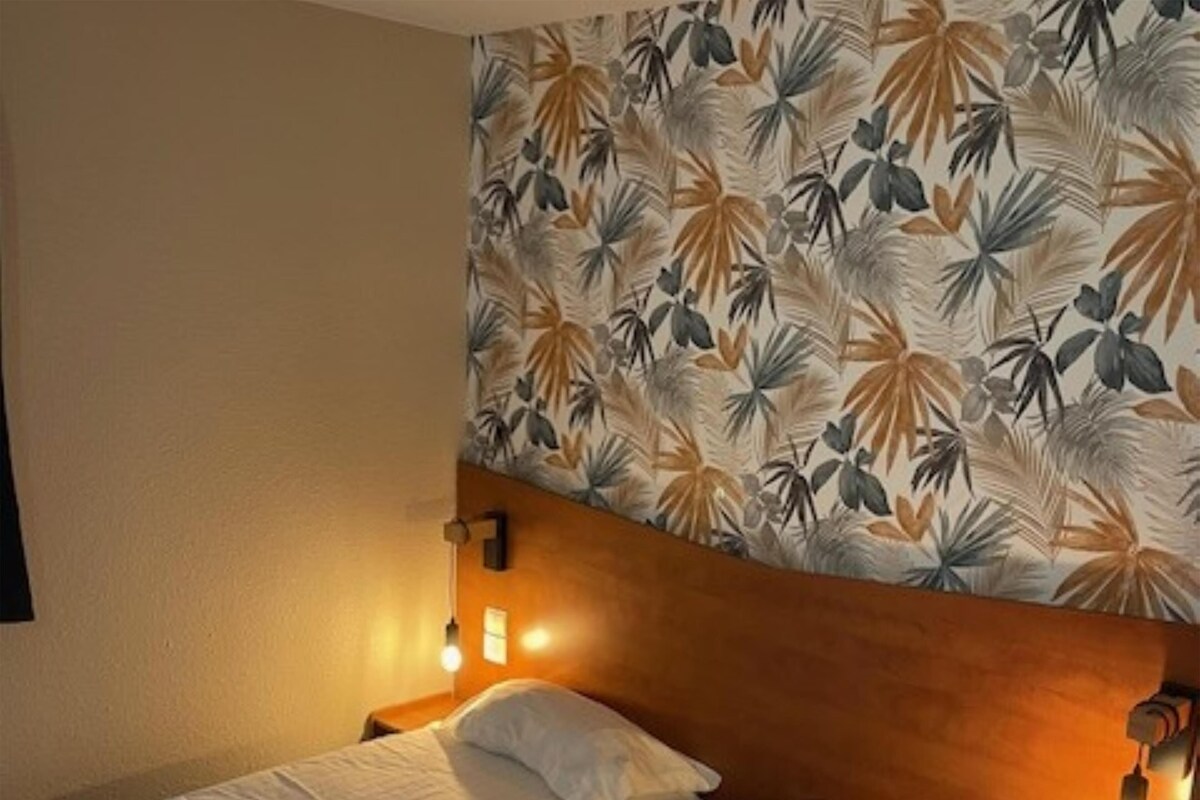 Une chambre d’hôtel avec deux lits simples, une tête de lit en bois et un mur orné d’un motif de feuilles tropicales.