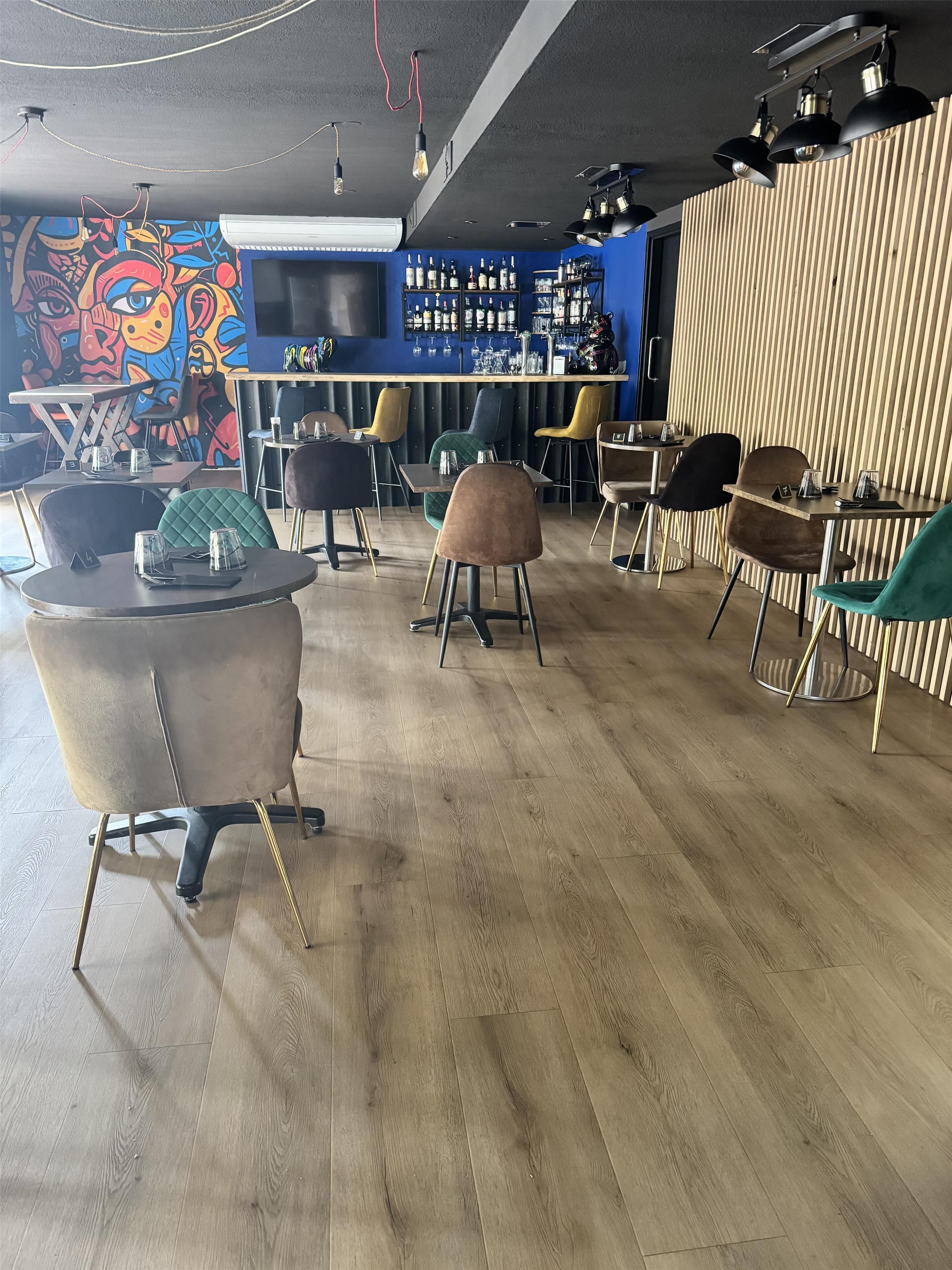 Una zona de bar con mostrador de madera, varios estantes llenos de botellas y un mural colorido en la pared.