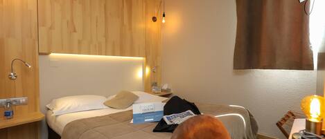 Quarto de hotel com cabeceira de madeira, cama com colcha bege, mesa de cabeceira com abajur e janela com cortina marrom.