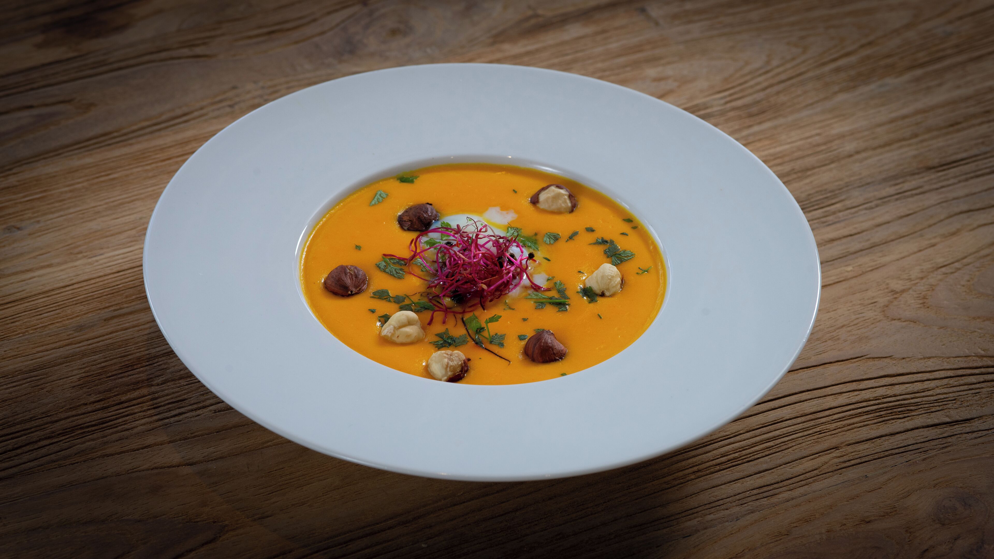 A white plate with a bowl of soup garnished with nuts, herbs, and a decorative element on a wooden table.