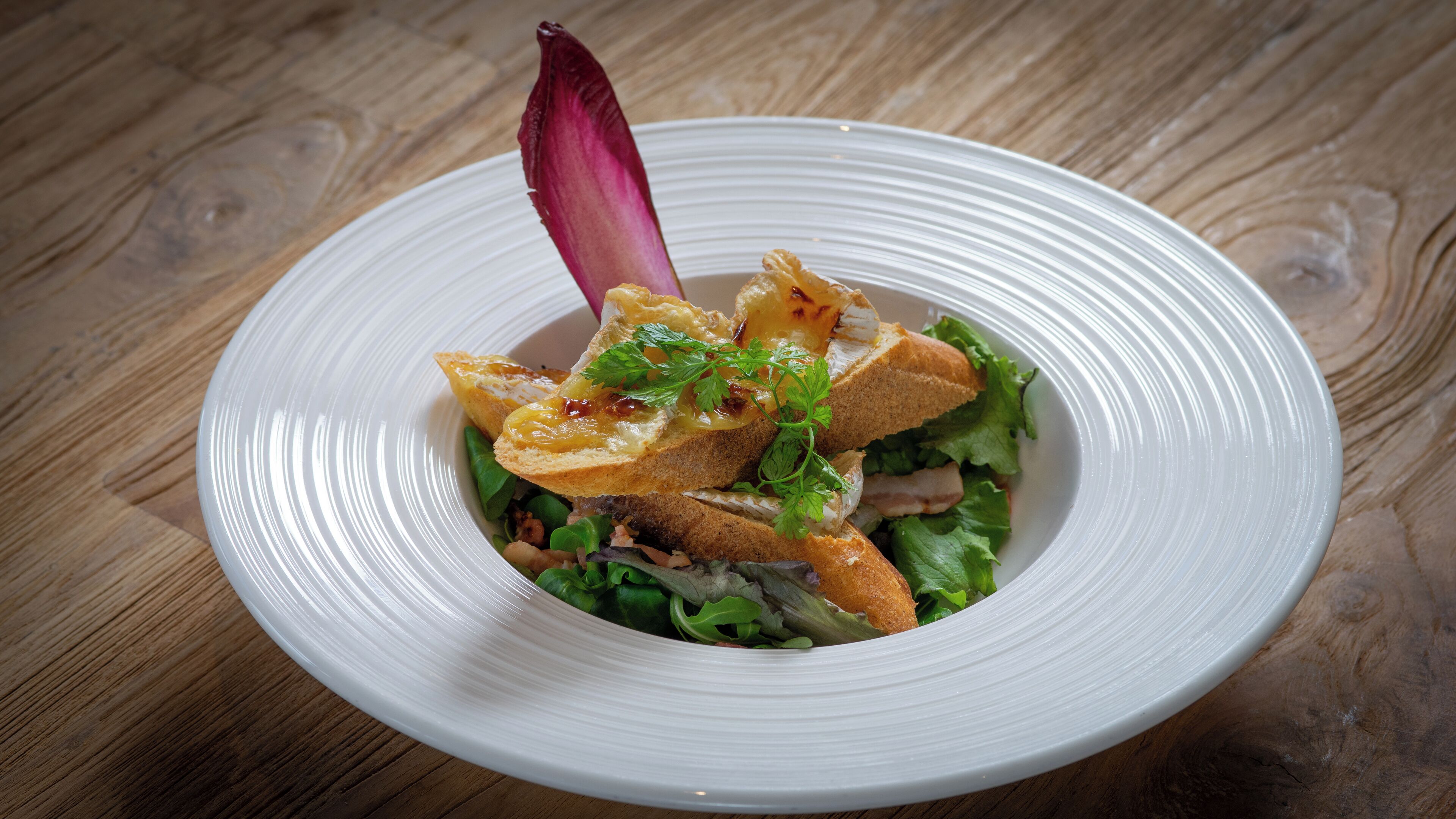 A white plate with a salad, bread, and a decorative purple element on a wooden table.