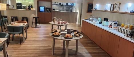 Un espace moderne dédié au petit-déjeuner, doté d’un sol en bois, d’un buffet central et d’un coin salon avec des tables et des chaises.