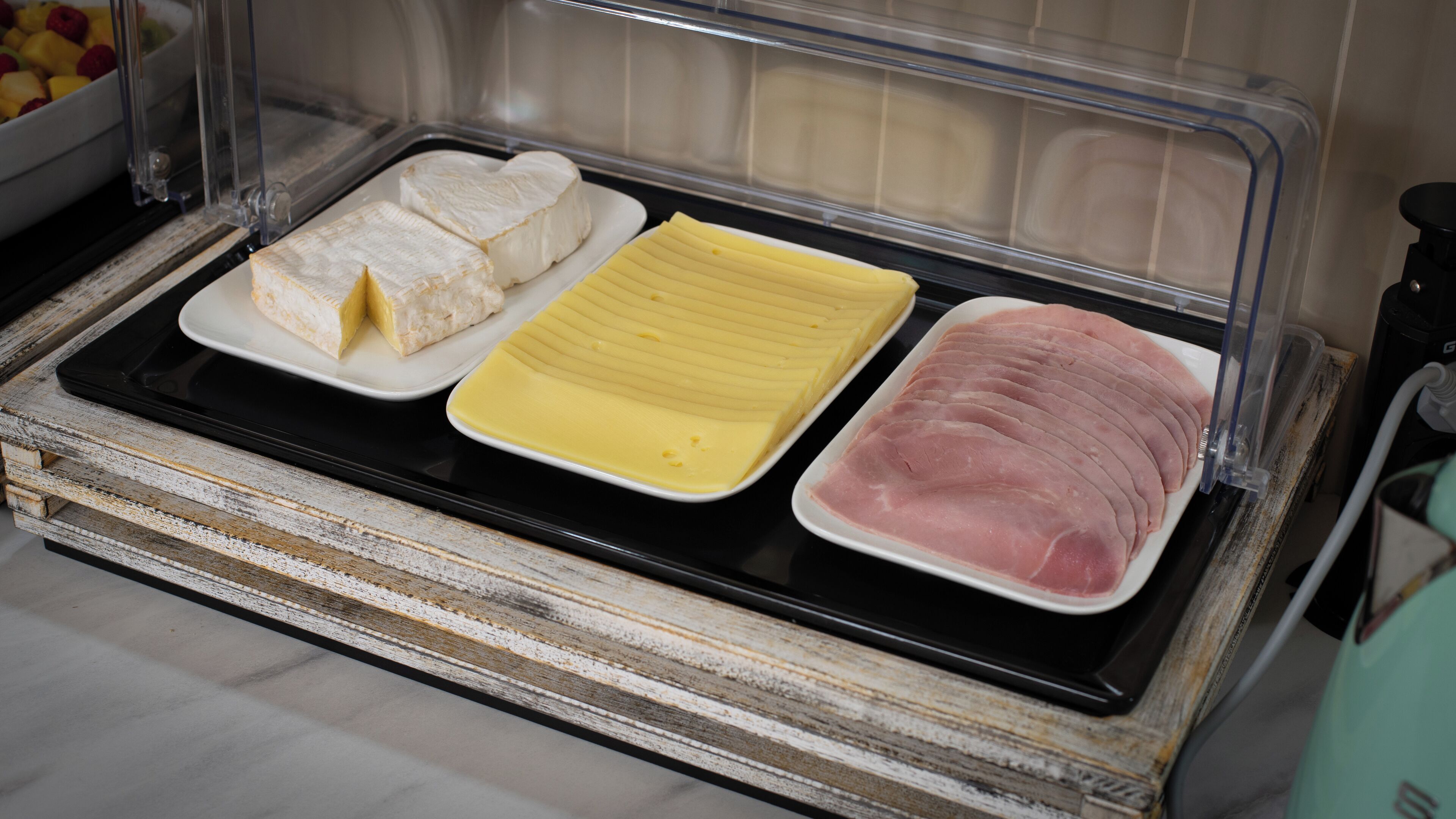A display of food items including cheese, ham, and a yellow spread on a black tray in a refrigerated display case.