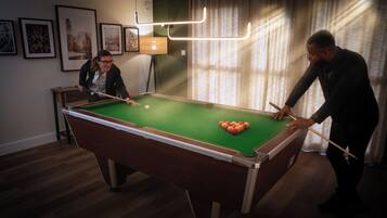 Une table de billard dans une pièce dont les murs sont ornés de tableaux encadrés et où se trouve une lampe.
