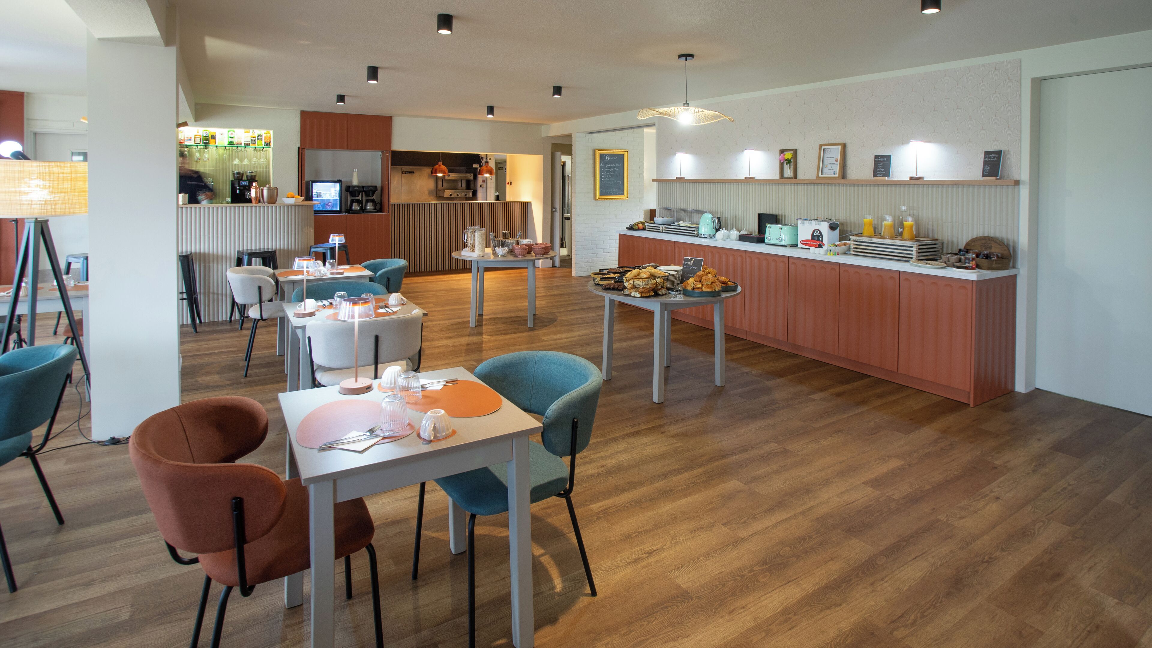 A modern hotel dining area with wooden flooring, a central island with a coffee machine, and a bar area with a chalkboard menu.