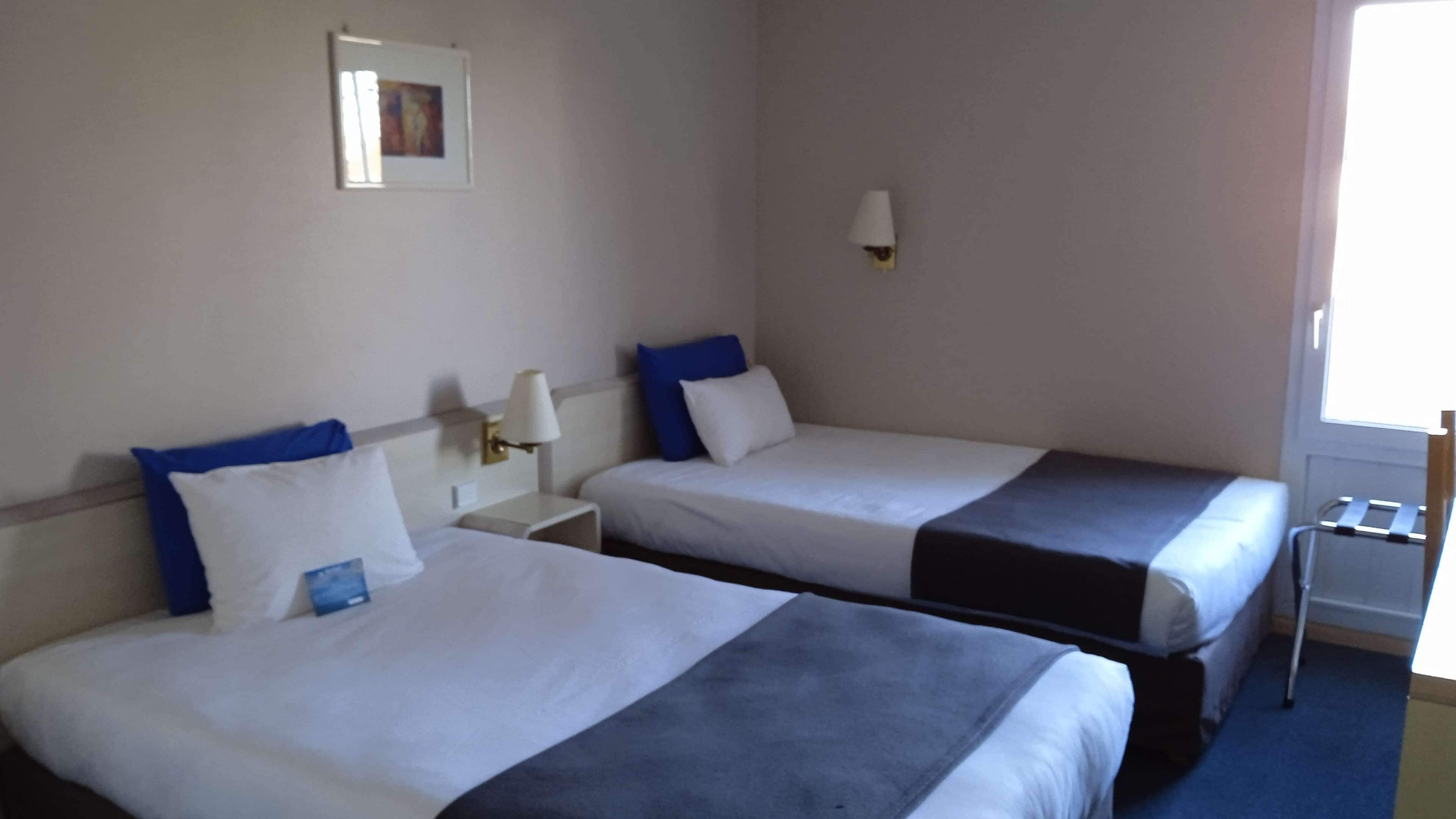 Habitació d'hotel amb dos llits, cada un amb roba de llit blanca i almoades blaves, una taula de nit amb una làmpada, i una finestra amb vista.