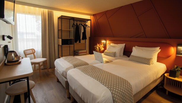 A hotel room with a large bed, a desk with a kettle, a chair, a wardrobe, and a bedside table with a lamp.