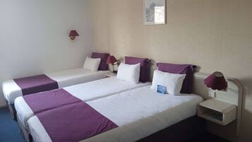 Habitación de hotel con dos camas, cada una con una colcha morada y almohadas blancas. Se observa un mesita de noche con una lámpara y una pequeña mesa con una tarjeta encima.