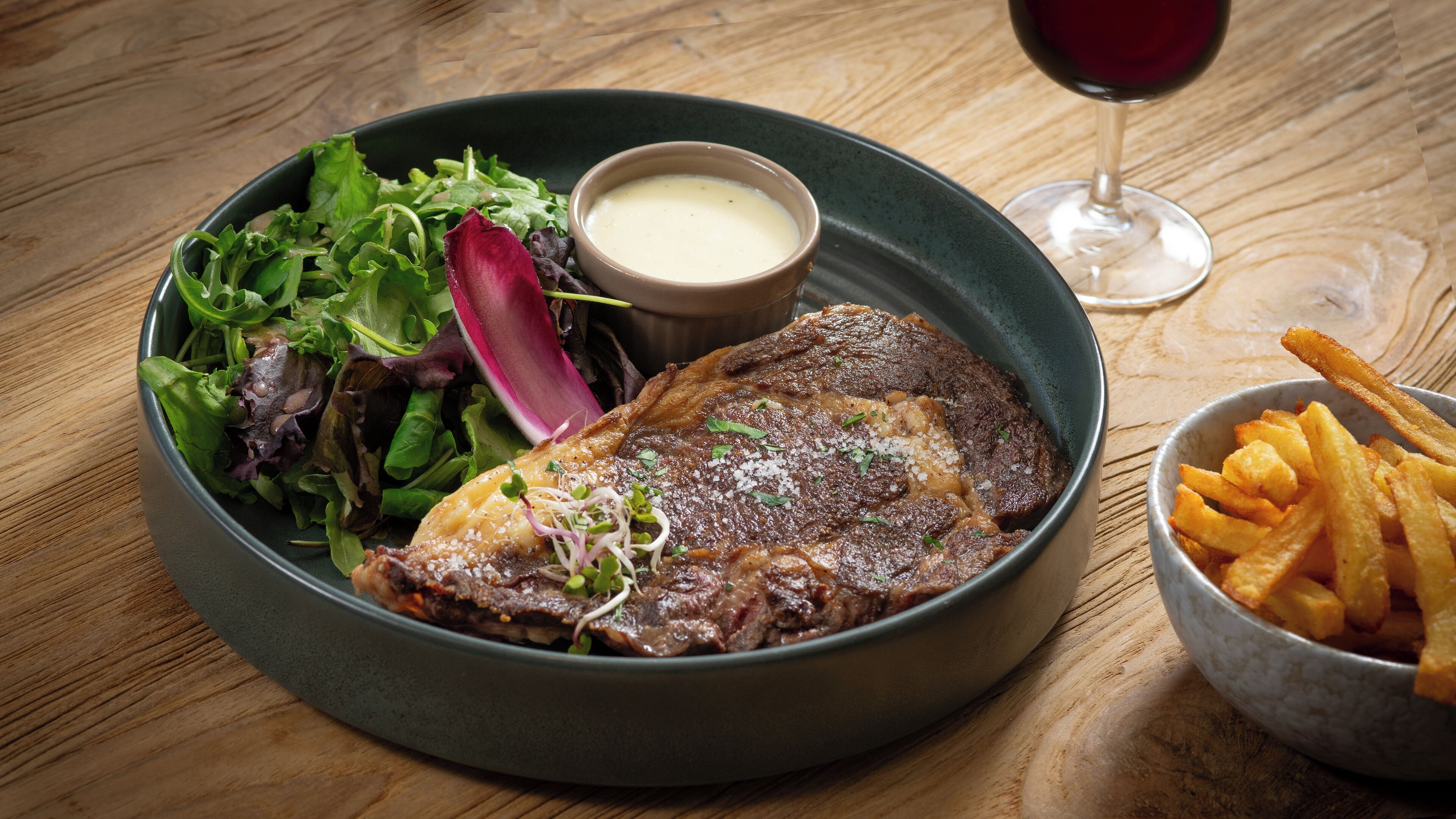 Une assiette comprenant un steak, une salade et des frites, accompagnée d’un verre de vin rouge.