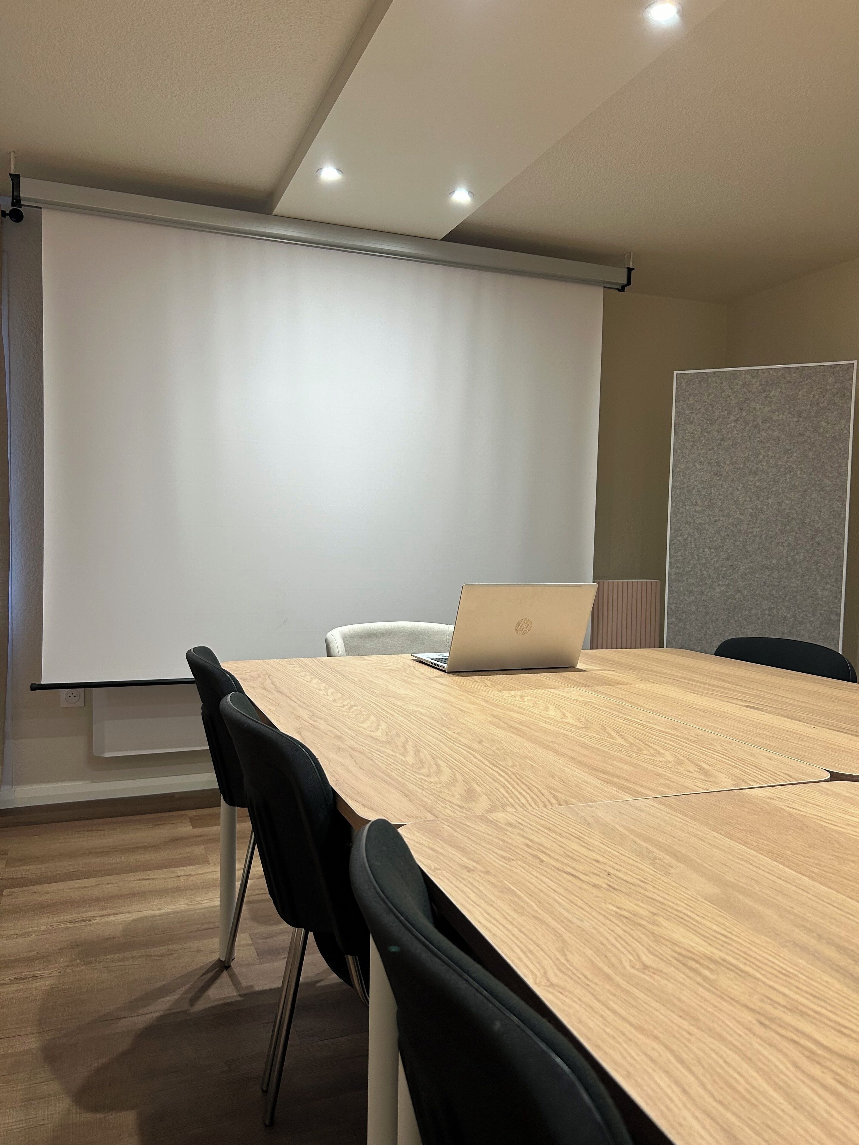 Una sala de conferencias con una mesa de madera, sillas negras, una computadora portátil, una pantalla de proyector y una pizarra.