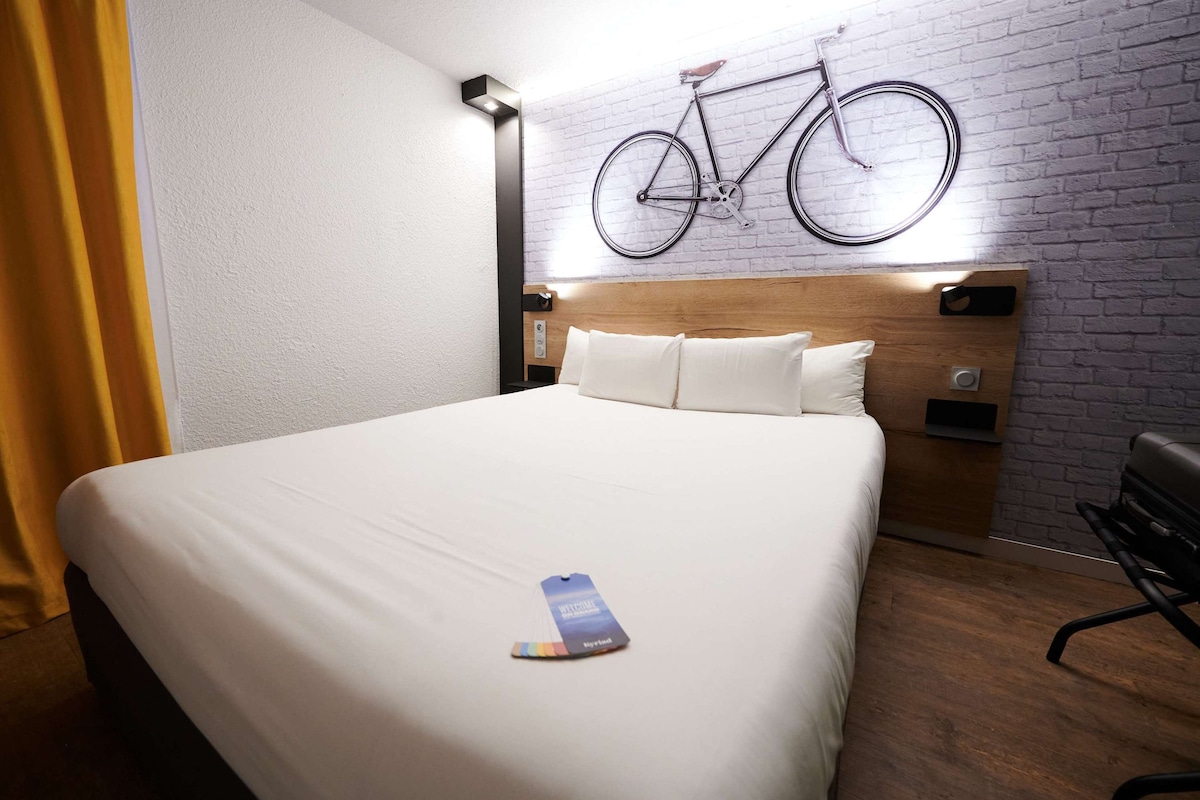 A neatly made bed with a white comforter and pillows, a wooden headboard, a wall-mounted bicycle, and a small nightstand with a lamp.