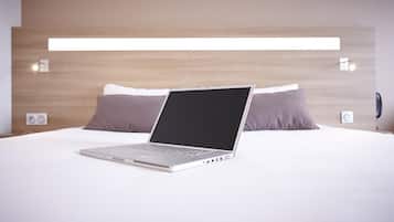 A modern hotel room with a laptop on the bed, a wooden headboard, and two wall-mounted speakers.