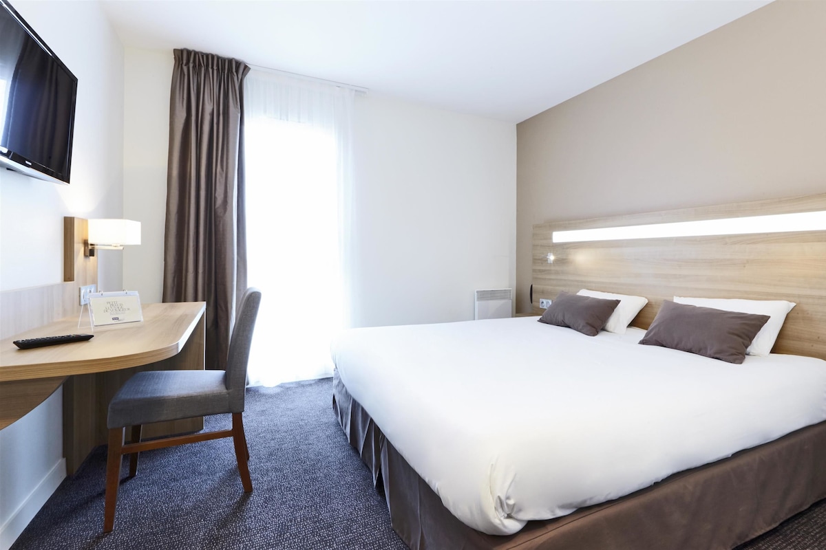 A hotel room with a bed, a desk with a chair, a TV mounted on the wall, and a window with curtains.