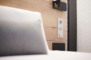A bed with a white pillow, a wooden headboard, and a power outlet on the wall.